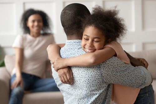 Child hugging a parent while the other parent is still on the couch.