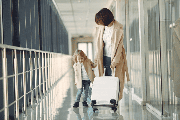 Woman at an airport, holding luggage in one hand, and a young girl’s hand in the other.